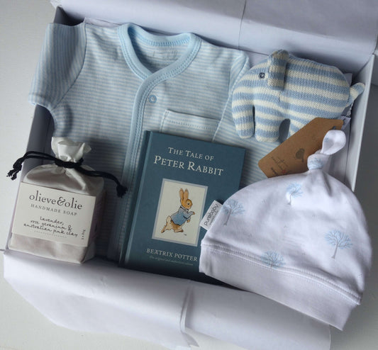 A baby gift set arranged in a white box, including a light blue striped baby romper, a blue and white striped plush elephant toy, a white beanie with blue tree prints, a book of The Tale of Peter Rabbit, and a small bag of olive&elie handmade soap.