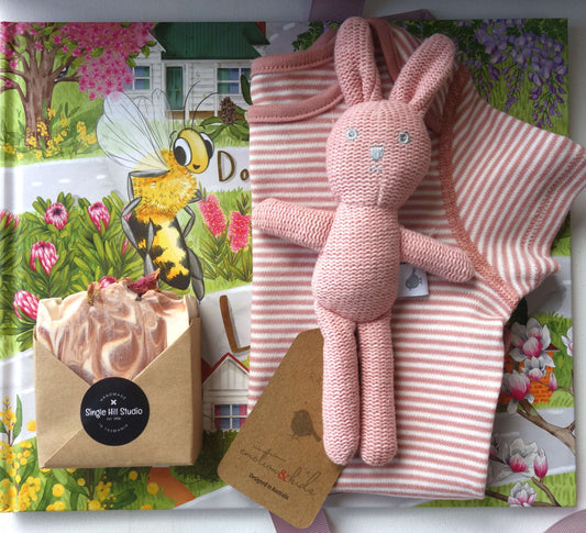 Baby girl gift hamper featuring a pink bodysuit, rabbit rattle, eco-friendly soap, and illustrated storybook.