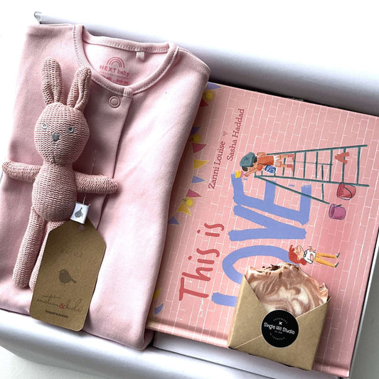 Welcome baby girl gifts including a pink bodysuit, toy rabbit, keepsake book, and eco-friendly soap.